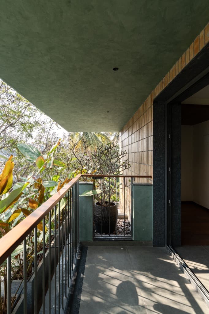 Cantilevered green balcony terrace with olive plaster soffit, potted frangipani, vertical brick cladding, and lush treetop canopy view