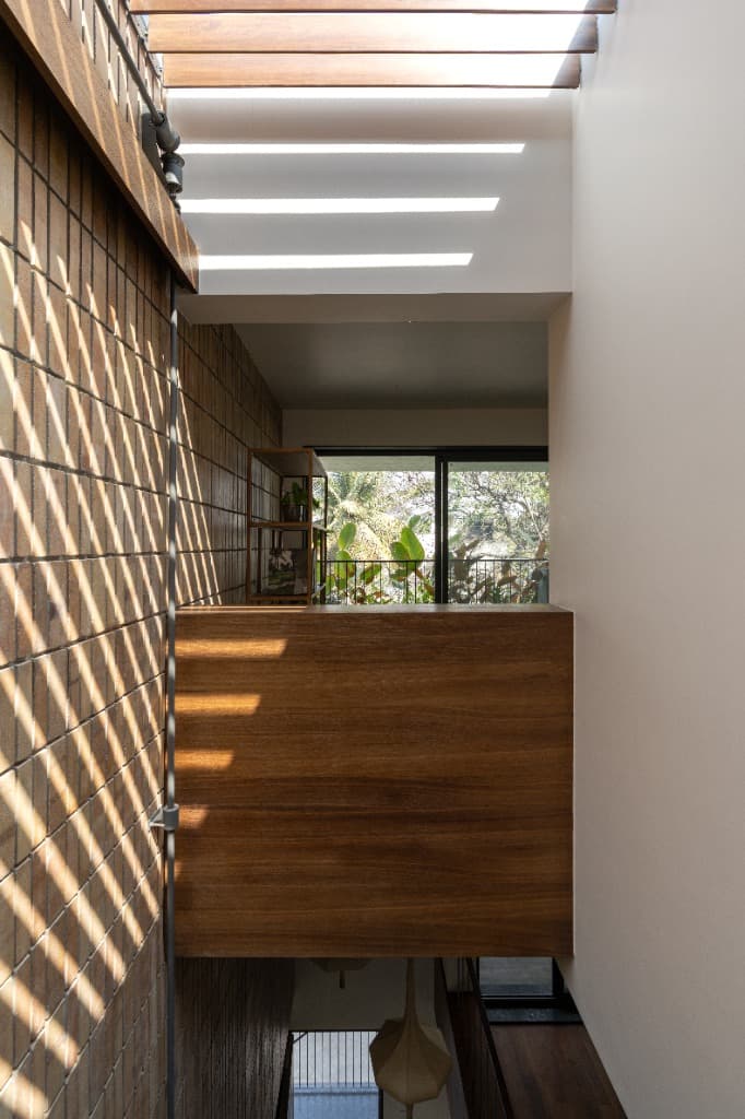Perspective down the lightwell court over solid teak bridge balustrade toward buff brick wall and pergola shadow bands