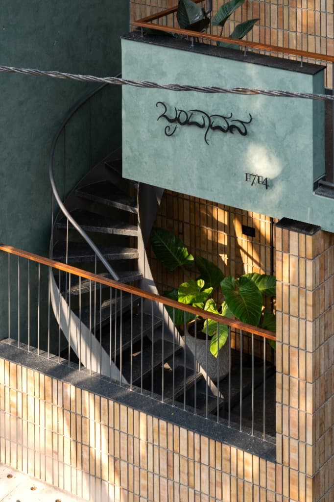 Close-up of the cantilevered sage green balcony corner with Kannada 'Brindavan' brass typography, house number 1714, and black granite spiral steps