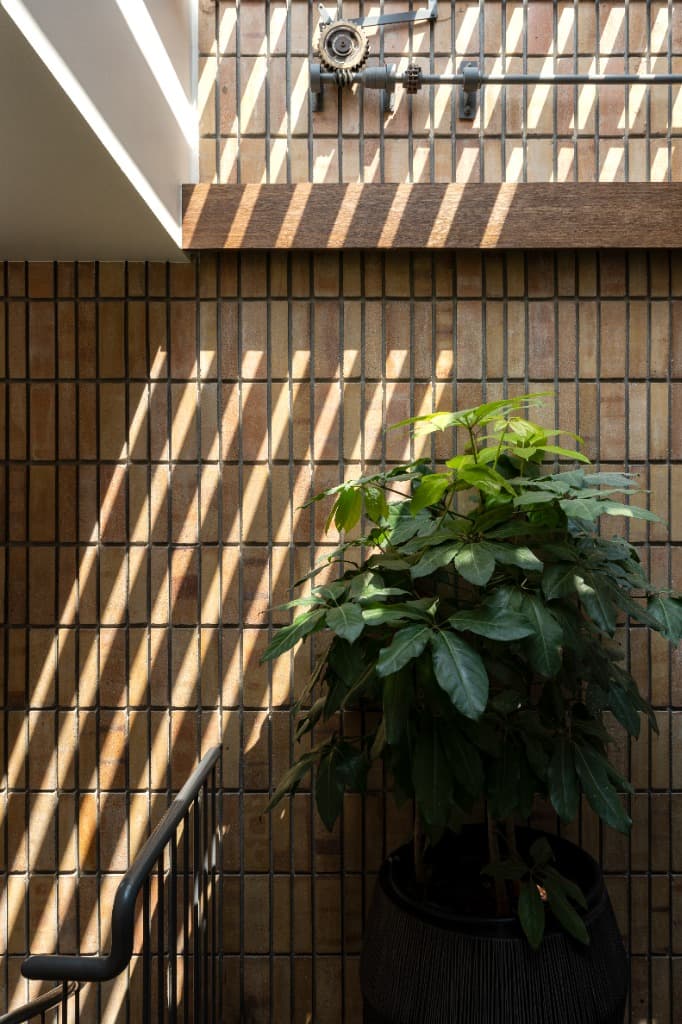 Stairwell lightwell showcasing vertical buff brick bond, potted indoor ficus, manual gear-lever mechanism, and linear skylight shadowplay
