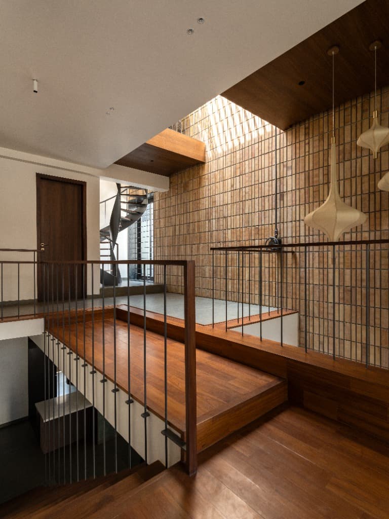 Upper level teak-lined bridge walkway overlooking the double-height lightwell court, sculptural cocoon pendants, and spiral stair landing