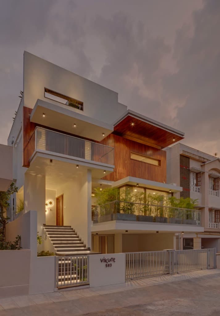 Three-quarter dusk perspective highlighting illuminated entrance steps, backlit wall sculpture, and cantilevered teak canopies
