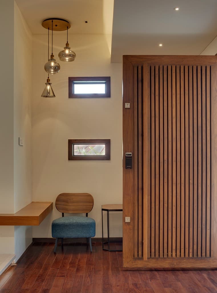 Foyer entrance anchored by an oversized solid teak pivot door with vertical fluting, brass drop pendants, and hardwood flooring